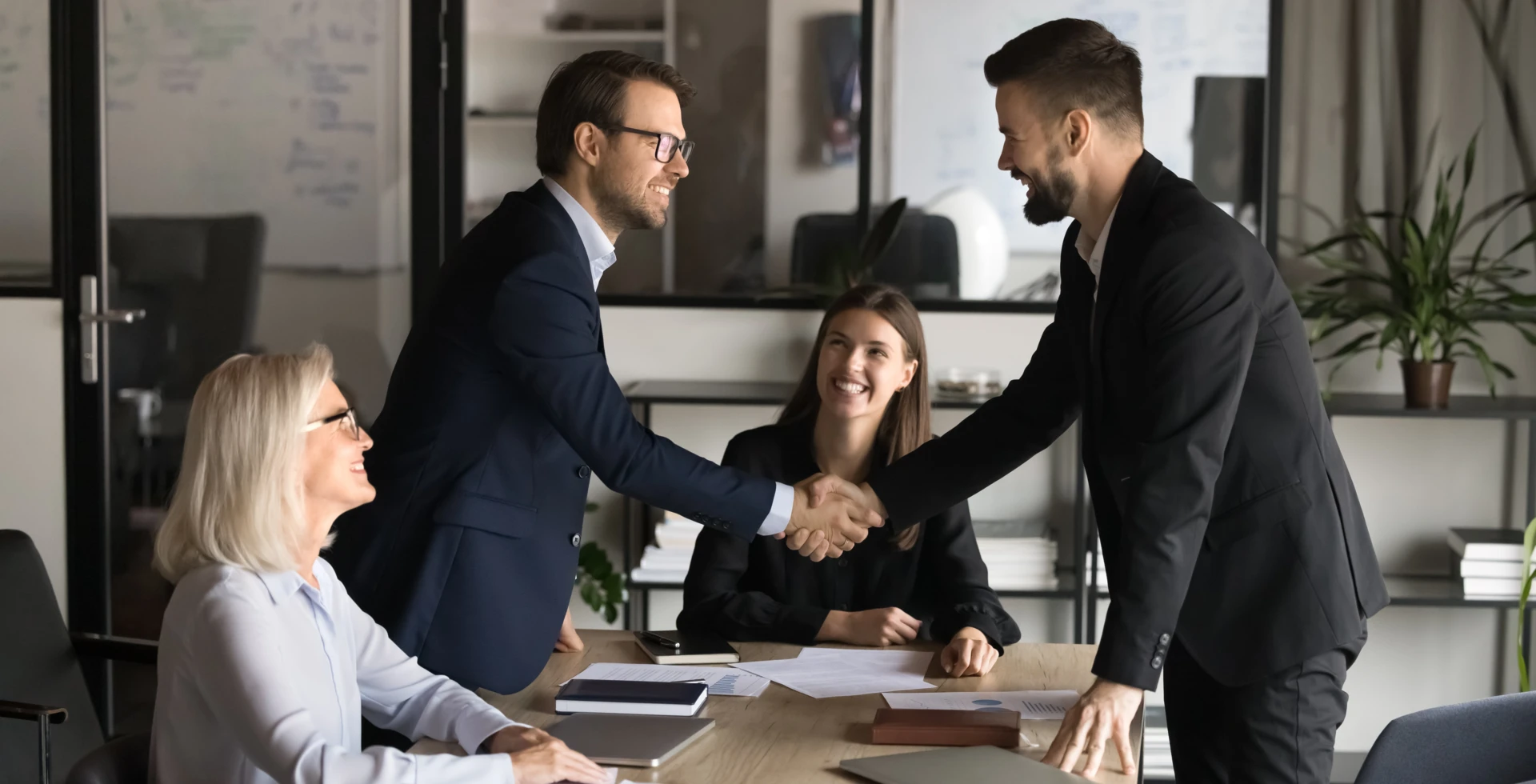 Grupa pracowników umysłowych w formalnych ubraniach siedząca wokół stołu w sali konferencyjnej. Omawiają pracę i współdziałają ze sobą.